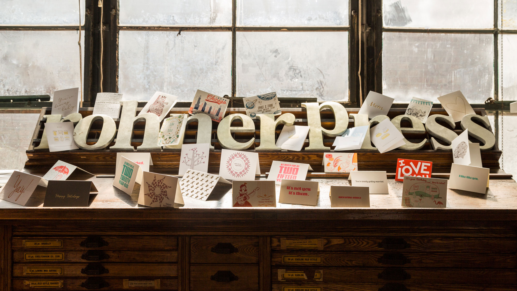 A wide shot of a weathered wooden desk with many small drawers, set against a large window. On the desk, large, cream-colored 3D letters spell out 'Rohner Press'. In front of and around these letters, numerous small greeting cards with various designs are displayed. The cards feature a mix of colors and patterns, some with text like 'LOVE' and 'TWO'. The window behind is large and has multiple panes, showing a bright, somewhat overcast sky outside.